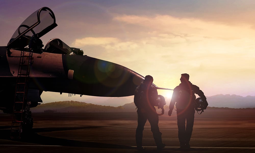 Two fighter pilots walk up to a jet, backlit by the setting sun