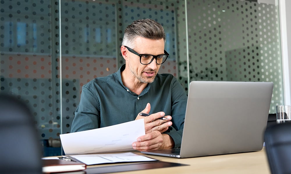 A man with gray hair and glasses talks on a video call at his desk. He