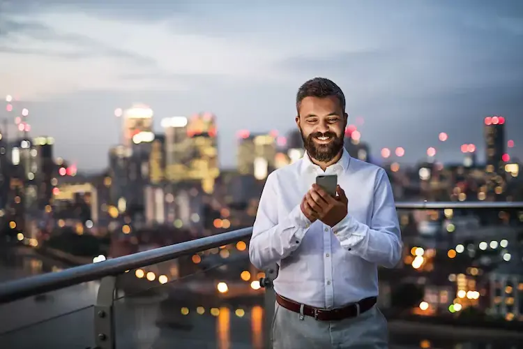 A man looking at his phone, standing on a balcony overlooking a downtown area. 