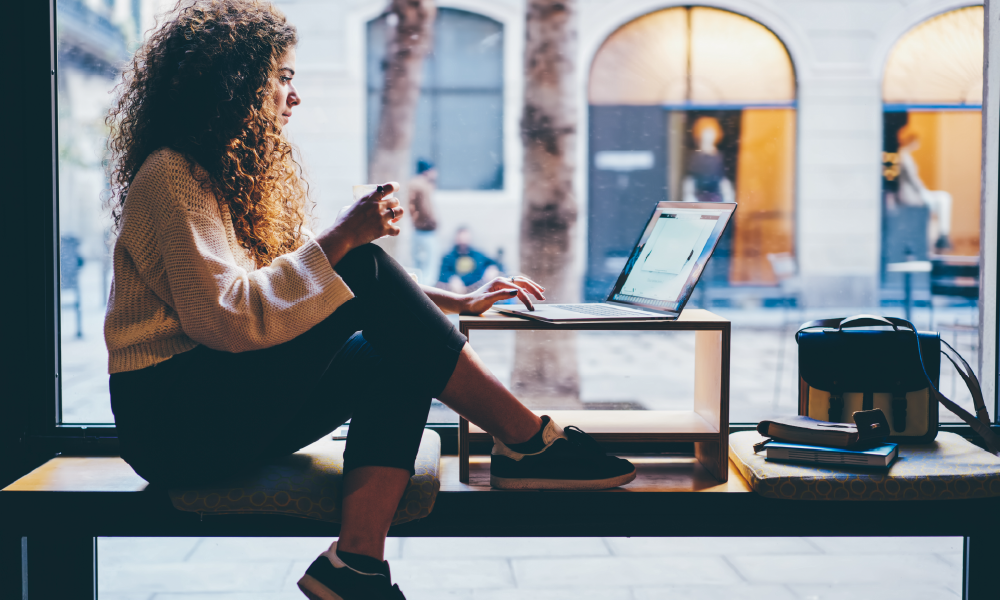 A woman sits in a coffee shop window working on her laptop.