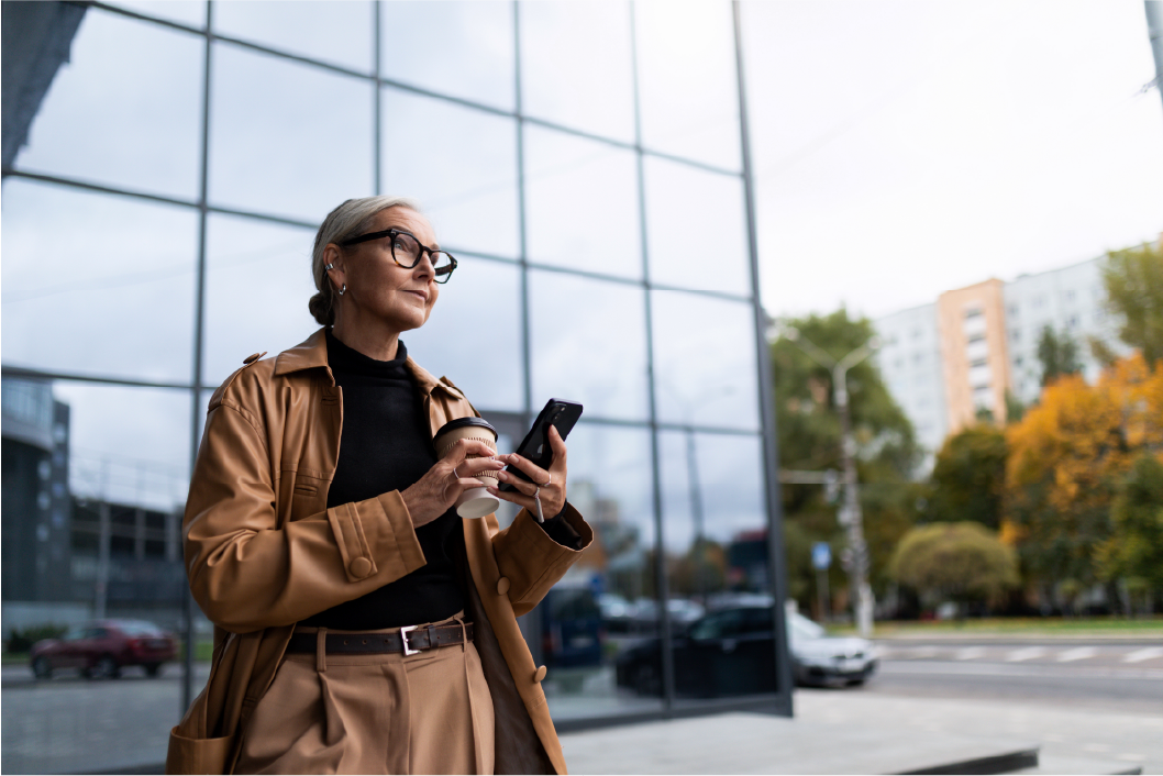 A photo of a woman in a brown trenchcoat, holding her phone and walking outdoors. 