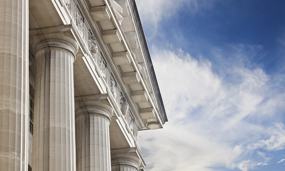 The side of a government building with marble columns, set against a blue sky with fluffy clouds