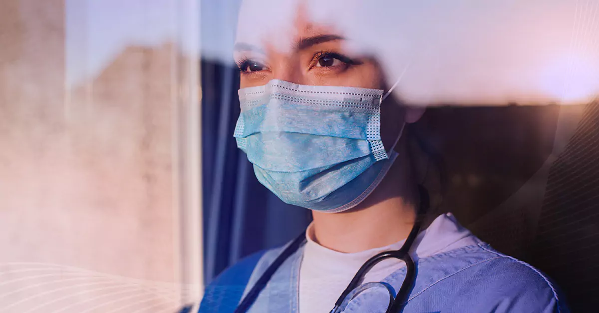 A woman wearing scrubs, a stethoscope, and a mask looks out the window