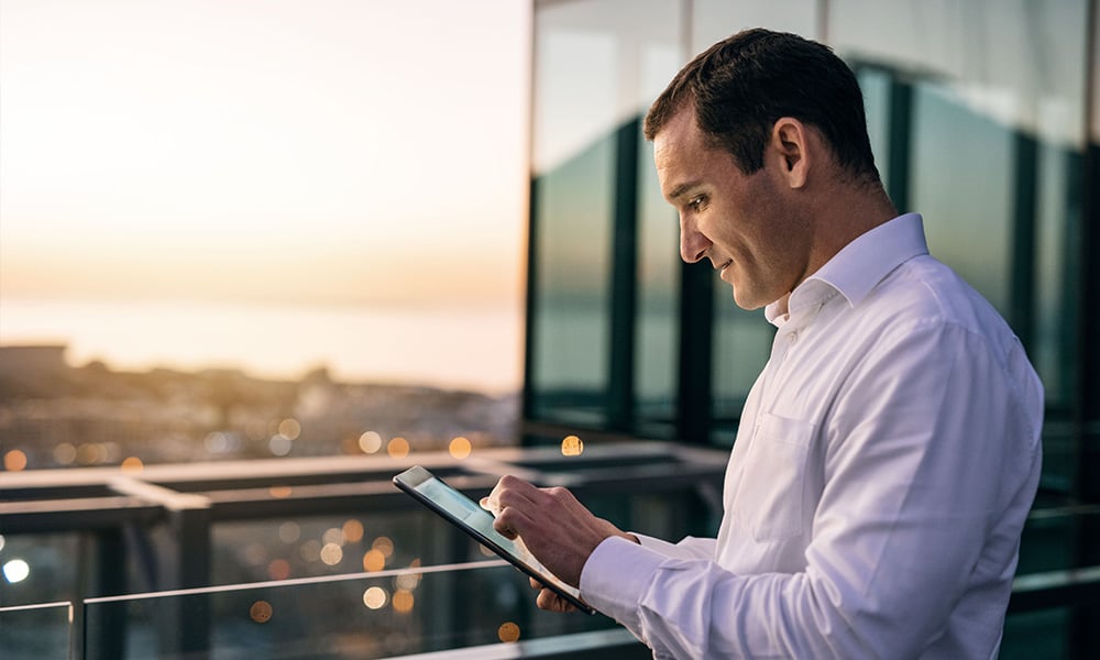 A man wearing a collared shirt uses a tablet while standing outside on a balcony, overlooking the city at sunset.